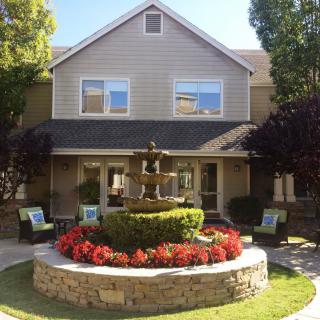 A two-story house with a beautifully landscaped garden featuring a central fountain surrounded by vibrant flowers.