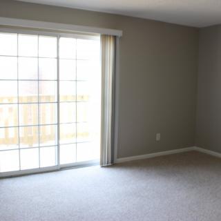 A sparsely decorated room with grey walls, featuring a sliding glass door that allows natural light to enter, and carpeted flooring.