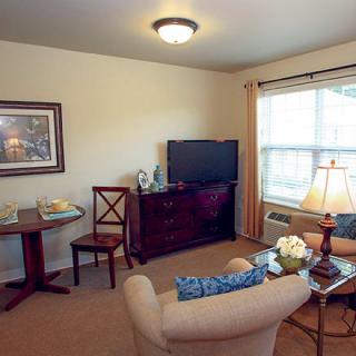 A cozy living room with comfortable seating, a television, and decorative elements, illuminated by natural light from a window.