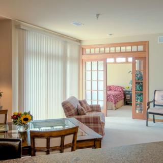 A cozy living room area with large windows, featuring comfortable furniture and a view into an adjacent bedroom.