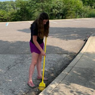 A young woman uses a yellow tool to clean the curb on a sunny day.