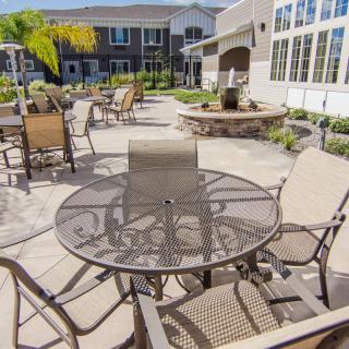 A sunny outdoor patio area with chairs and tables, featuring a fire pit and surrounded by a residential building.