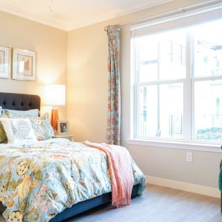 A well-lit bedroom featuring a comfortable bed with floral bedding, a bedside lamp, and large window.