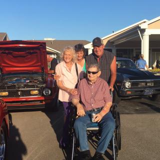 A group of people posing outdoors in front of cars, with one person in a wheelchair.