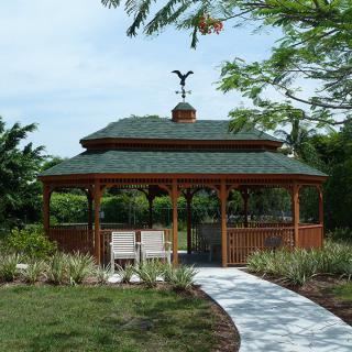 A wooden gazebo with a green roof located in a garden, surrounded by trees, with a pathway leading up to it.