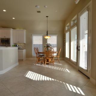 A bright kitchen and dining area with natural light streaming through blinds, featuring a wooden table and chairs on tiled flooring.
