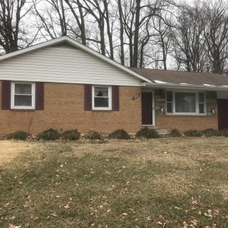 A single-story ranch-style brick house with a front yard and surrounding trees.