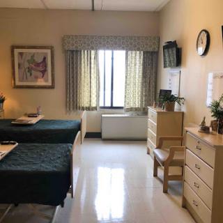 A well-lit hospital room featuring two patient beds, a window with curtains, and a simple dresser with decorative items.