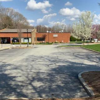 An empty parking lot in front of a brick building surrounded by trees and under a partly cloudy sky.