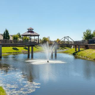 A serene view of a park featuring a fountain, gazebo, and a bridge over the calm waters.