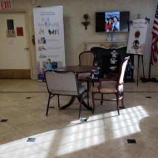 A cozy lobby area featuring a table and chairs, with a television displaying images, banners on display, and natural light illuminating the space.