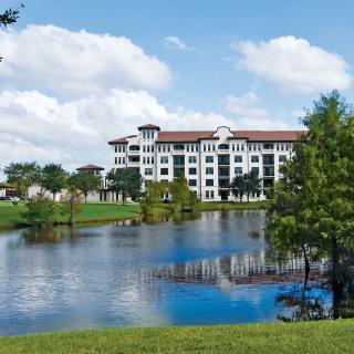 A scenic view of a building by a lake surrounded by trees and under a blue sky with clouds.