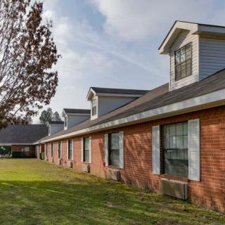 A long brick building with multiple windows and a sloped roof sits beside a green lawn and tree on a clear day.