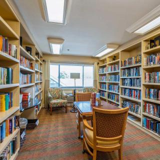A cozy library with bookshelves, a table, and chairs, lit by natural light from the window.