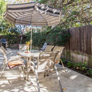 A sunny outdoor patio with a table, chairs, and an umbrella in a garden setting.
