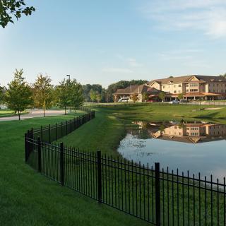A scenic view of a residential area with a pond, surrounded by grass, trees, and a black metal fence.