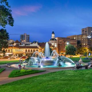A beautifully lit fountain in a city park at dusk, with buildings and lights in the background.