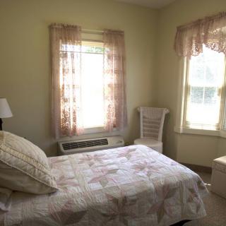 A cozy bedroom with natural light streaming through sheer curtains, featuring a neatly made bed, a bedside lamp, and a chair by the window.