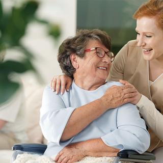 A caregiver warmly embracing an elderly woman, both smiling and sharing a moment of joy indoors.