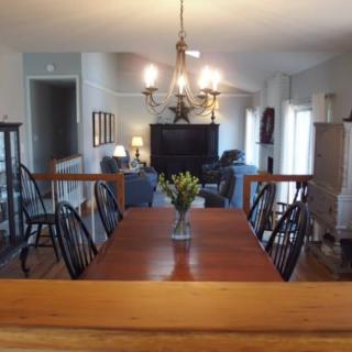 A modern dining area with a wooden table and black chairs, adorned by a chandelier, opens to a living room.