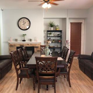 A cozy dining and living area featuring a wooden table with chairs, sofas, a wall clock, shelves, and a ceiling fan on hardwood floors.