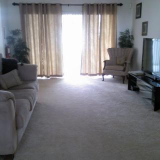 A spacious living room with a sofa, armchair, and a television, featuring large curtains that allow natural light to filter in.