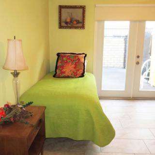 A small bedroom with a single bed covered in green bedding, featuring a colorful pillow, a side table with a lamp, and large windows letting in natural light.