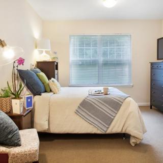 A cozy bedroom with a neatly made bed, colorful cushions, and a large window letting in natural light.