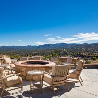 A scenic outdoor patio with wooden chairs surrounding a fire pit, overlooking a mountainous landscape under a clear blue sky.