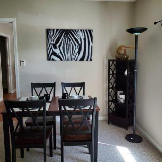 A dining area with a wooden table and four chairs, featuring modern artwork on the wall and a tall floor lamp beside a shelving unit.