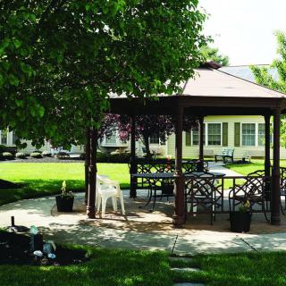 A peaceful gazebo with outdoor seating on a sunny patio surrounded by a lush green lawn and trees.