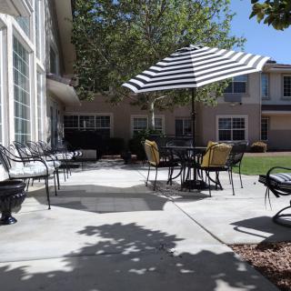 A sunny patio area with tables, chairs, and a striped umbrella surrounded by a garden and buildings.