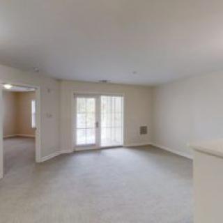 A spacious, empty living room with neutral tones, featuring carpet flooring and large windows letting in natural light.
