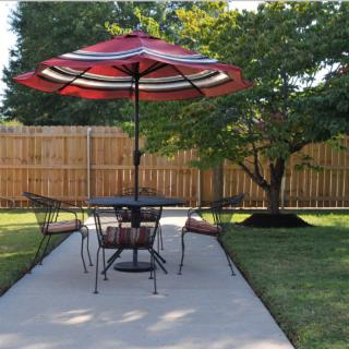 A patio with a table and chairs under a red umbrella, surrounded by a lush garden.