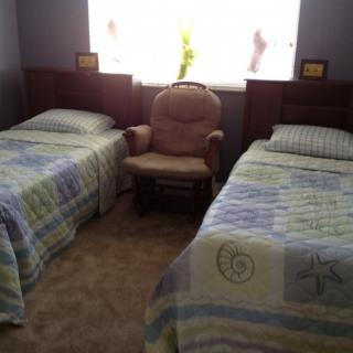 A cozy bedroom features two twin beds with patterned quilts and a comfortable chair between them, illuminated by light from the window.