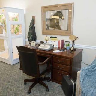 An office setup featuring a wooden desk with a typewriter, telephone, and lamp, accompanied by an office chair. A framed artwork hangs above the desk.