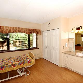 A well-lit bedroom featuring a hospital-style bed, large windows, and a simple wooden floor.