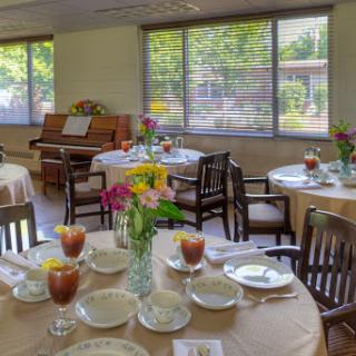 A cozy dining area set with tables adorned with flowers and refreshments, featuring natural light streaming through large windows.