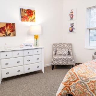 A bright and cozy bedroom with a white dresser, patterned chair, colorful bedding, and decorative artwork.