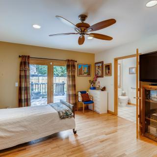 A cozy bedroom with wood flooring, a ceiling fan, and large windows overlooking the deck. The room features a neatly made bed, colorful curtains, and artwork on the walls. A bathroom is visible through an open doorway.