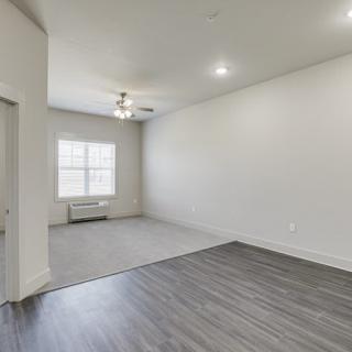 A modern, empty room with hardwood flooring and carpeted area, featuring white walls, a window, and ceiling lighting.