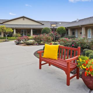 A tranquil outdoor area with a red bench and yellow cushion, surrounded by flowering plants and a well-maintained garden in front of a building.