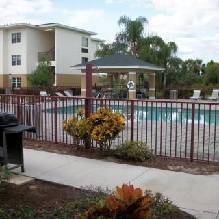 An outdoor area with an apartment complex, featuring a swimming pool, grill, and palm trees.