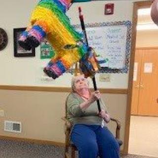 An elderly woman playfully hits a colorful piata indoors.