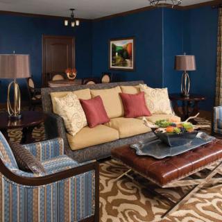 A stylish living room with blue walls, featuring a comfortable sofa and armchair, accented by red and beige cushions. A patterned rug complements the furniture, while lamps and artwork add decorative touches.