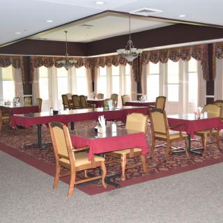 A spacious dining room with several tables and chairs, adorned with red tablecloths and surrounded by large windows with elegant curtains.