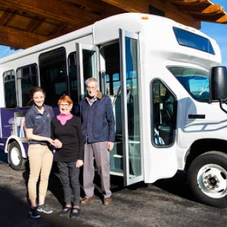 A group of three people stands in front of a new community bus, showcasing partnership and local transportation efforts.