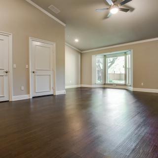 A spacious, empty room with hardwood flooring and a ceiling fan, illuminated by natural light from large windows.