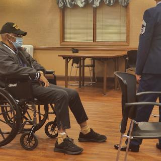 An elderly man in a wheelchair wearing a mask, seated indoors as a healthcare professional engages with him.