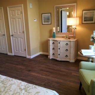 A cozy bedroom with wooden flooring, featuring a dresser, lamp, and artwork on the wall.
